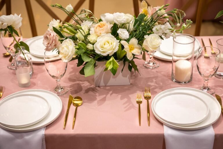 A beautifully arranged wedding table with a floral centerpiece and golden cutlery on a pink tablecloth.